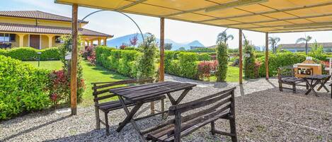 Un patio al aire libre con bancos y mesas de madera, una zona de barbacoa y un toldo amarillo.