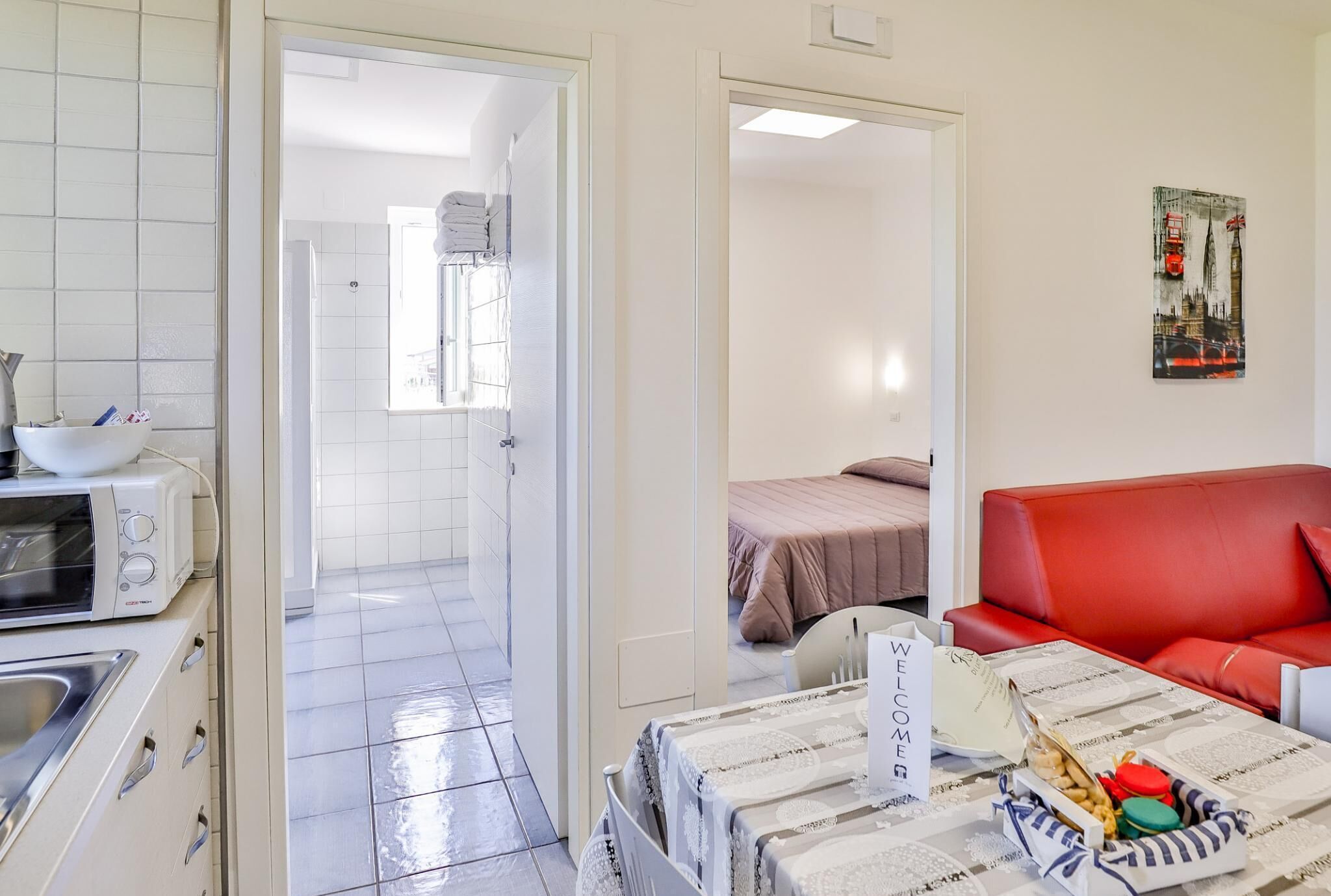 Quarto de hotel com cozinha compacta, cama com colcha rosa, sofá de couro vermelho e placa de boas-vindas.