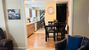A living room with a blue armchair, a dining area with a table and chairs, and a kitchen in the background. - Village House at Valley Village (Irwin)