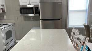 Una cocina con armarios blancos, un refrigerador de acero inoxidable, un microondas y una estufa. Hay una isla grande con encimera blanca y una silla con asiento de mimbre.