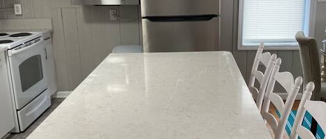 A kitchen with white cabinets, a stainless steel refrigerator, a microwave, and a stove. There is a large island with a white countertop and a chair with a woven seat.