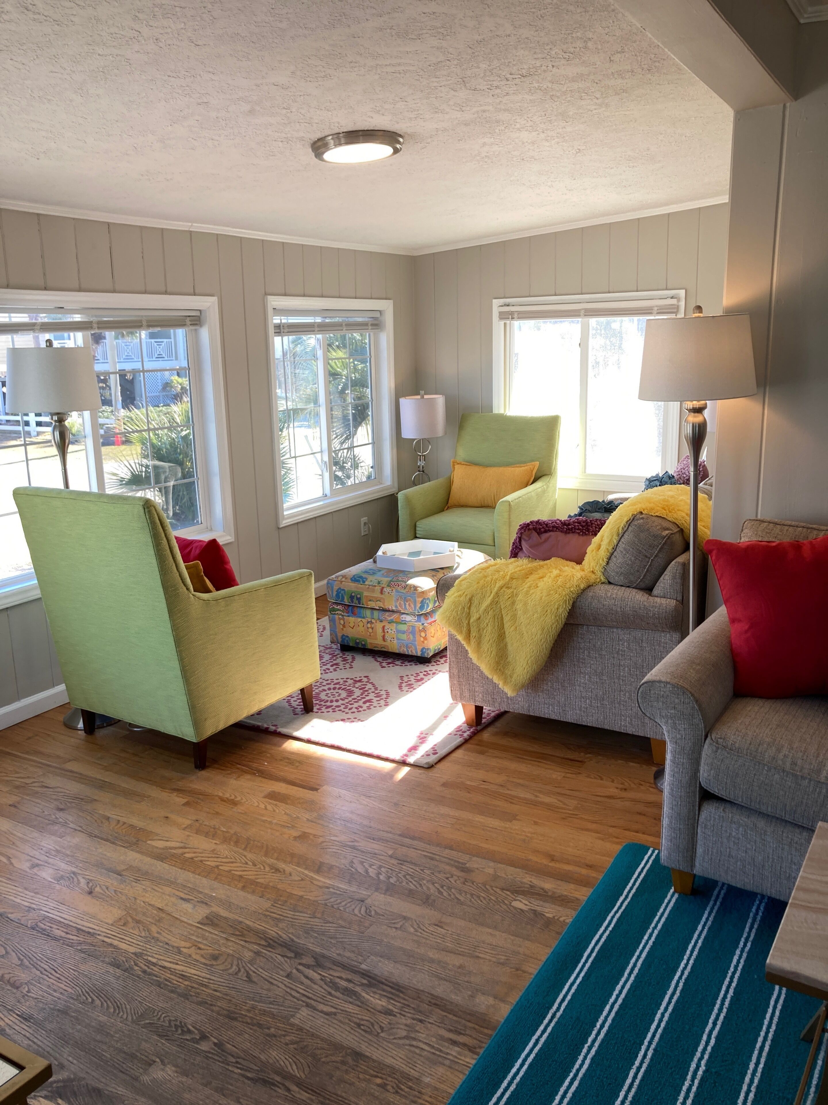 A living room with a green armchair, a grey sofa, and a yellow armchair. There are two windows with blinds, a lamp, and a patterned rug.