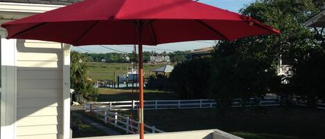 A red umbrella on a porch with a view of a white fence and greenery in the background.