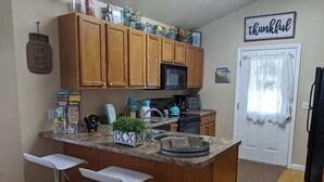 Una cocina con armarios de madera, encimera de granito, microondas y refrigerador. Hay dos taburetes blancos y una pequeña isla con una planta. Un letrero dice 'agradecido' en la pared.