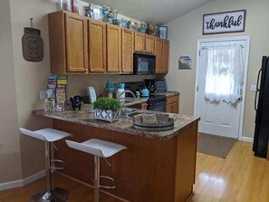 A kitchen with wooden cabinets, a granite countertop, a microwave, and a refrigerator. There are two white bar stools and a small island with a plant. A 'thankful' sign is on the wall. - The Southern Comfort (Pigeon Forge)