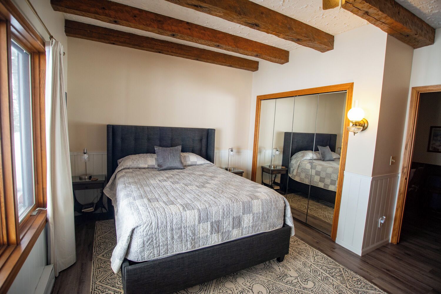 Camera da letto con un grande letto, travi in legno, uno specchio e un comodino con una lampada.