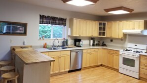A kitchen with wooden cabinets, a dishwasher, a stove, and a window with a view of greenery. - Your Home Away from Home in the Heart of the Kenai Peninsula (Soldotna)