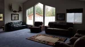 A spacious living room with a high ceiling, large windows, and a ceiling fan. - Your Home Away from Home in the Heart of the Kenai Peninsula (Soldotna)