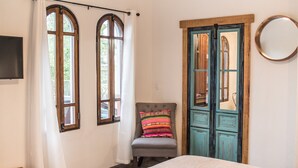 A bedroom with a bed, a chair with a colorful pillow, a wooden door, and a mirror on the wall. - Loreto80-NEW Studio Nopal, in downtown by beach (Loreto)