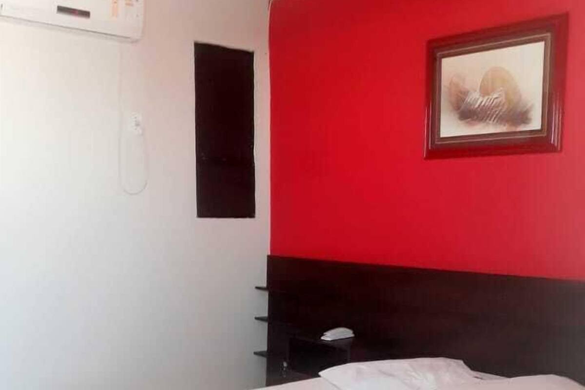 A hotel room with a bed, a nightstand, a red accent wall, a framed picture, and an air conditioning unit.