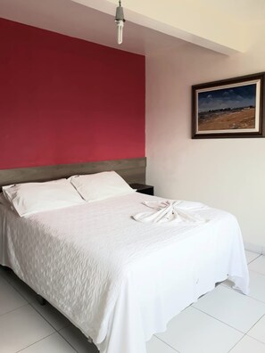 A neatly made bed with white linens and a towel, a red accent wall, and a framed picture on the wall. - Kris Hotel (Petrolina)