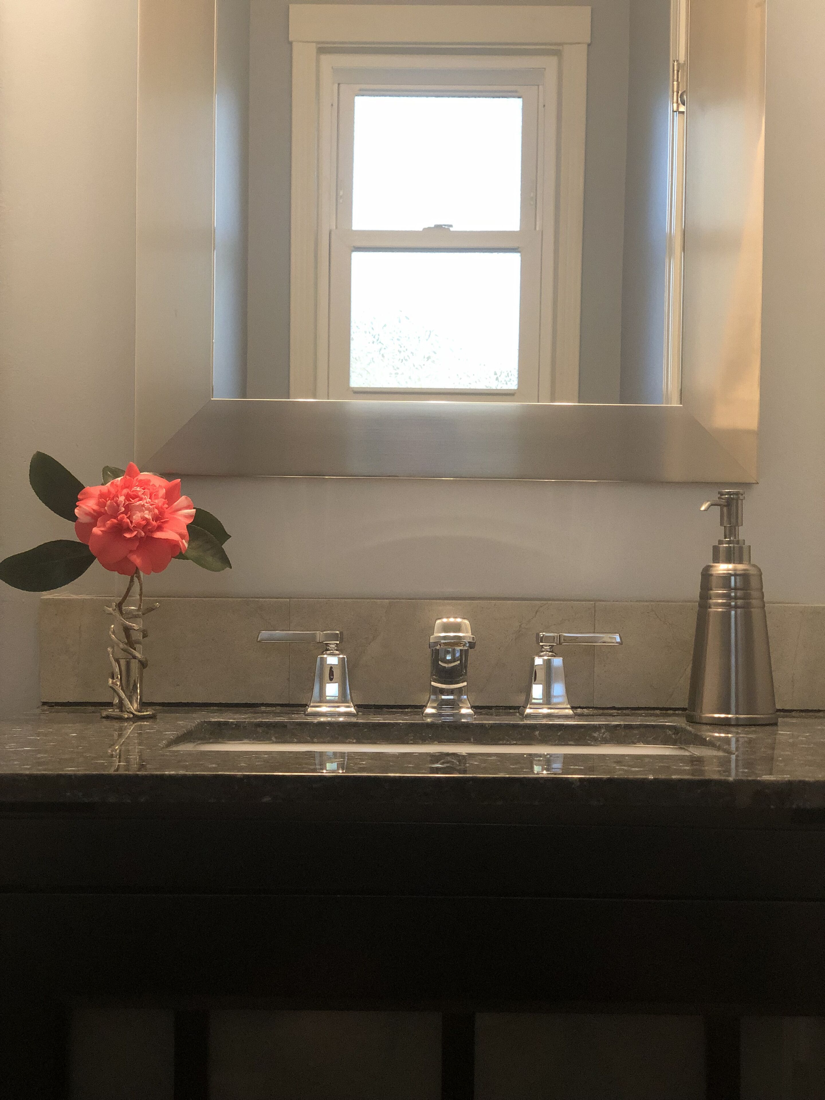 Un baño con lavamanos, tres grifos, dispensador de jabón y una flor en un florero.