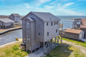 A waterfront property with a two-story house elevated on stilts, a dock, and a small boat. - Soundfront Dog Friendly Home w/ Hot Tub, Game Room, Private Boat Dock, Kayaks (Avon)