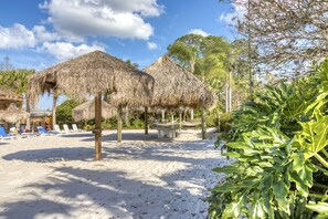 Relax in a serene beach setting with thatched-roof structures, lounge chairs, and a central seating area surrounded by palm trees and tropical plants.