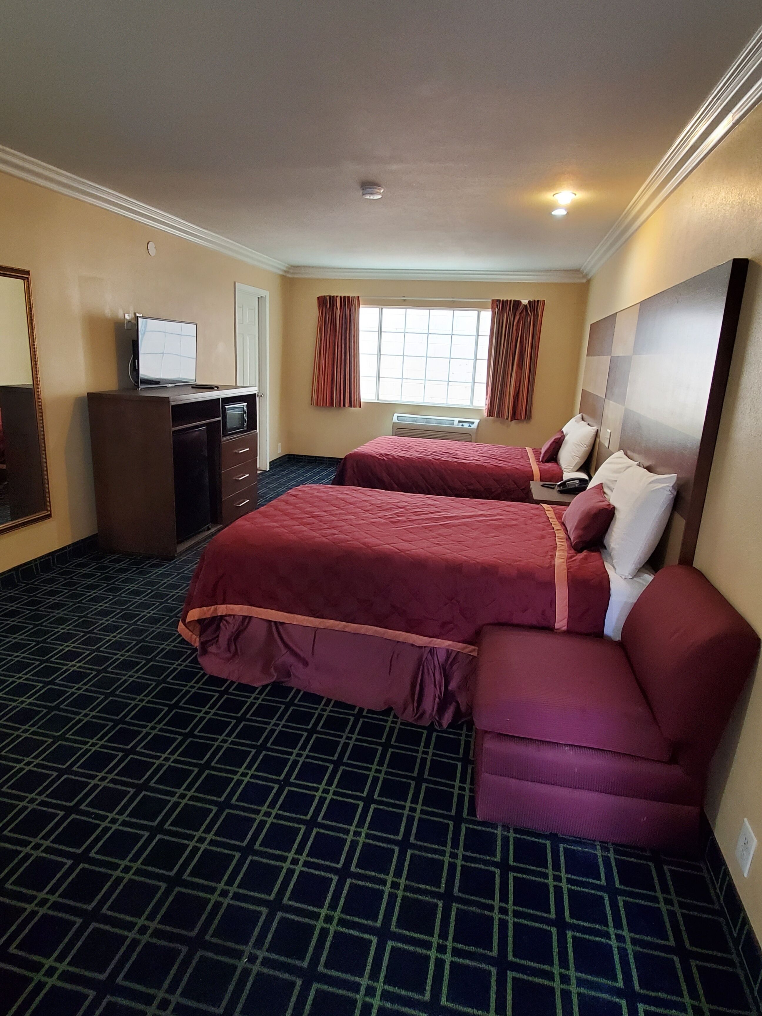A hotel room with two beds, a desk with a computer, a window with curtains, and a wall-mounted TV.