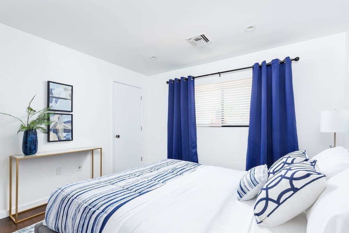 A bedroom with a bed, blue curtains, a bedside table, a lamp, and a wall-mounted picture.