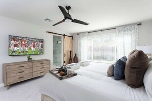 Un dormitorio con cama, televisor, ventilador de techo, cómoda y ventana con persianas.