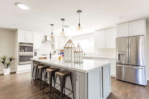Una cocina moderna con una isla central, taburetes de bar, electrodomésticos de acero inoxidable, iluminación colgante y una planta en maceta.