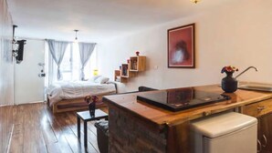 Een compacte kamer met een bed, een kleine keukenhoek, een houten eettafel en een enkele stoel.