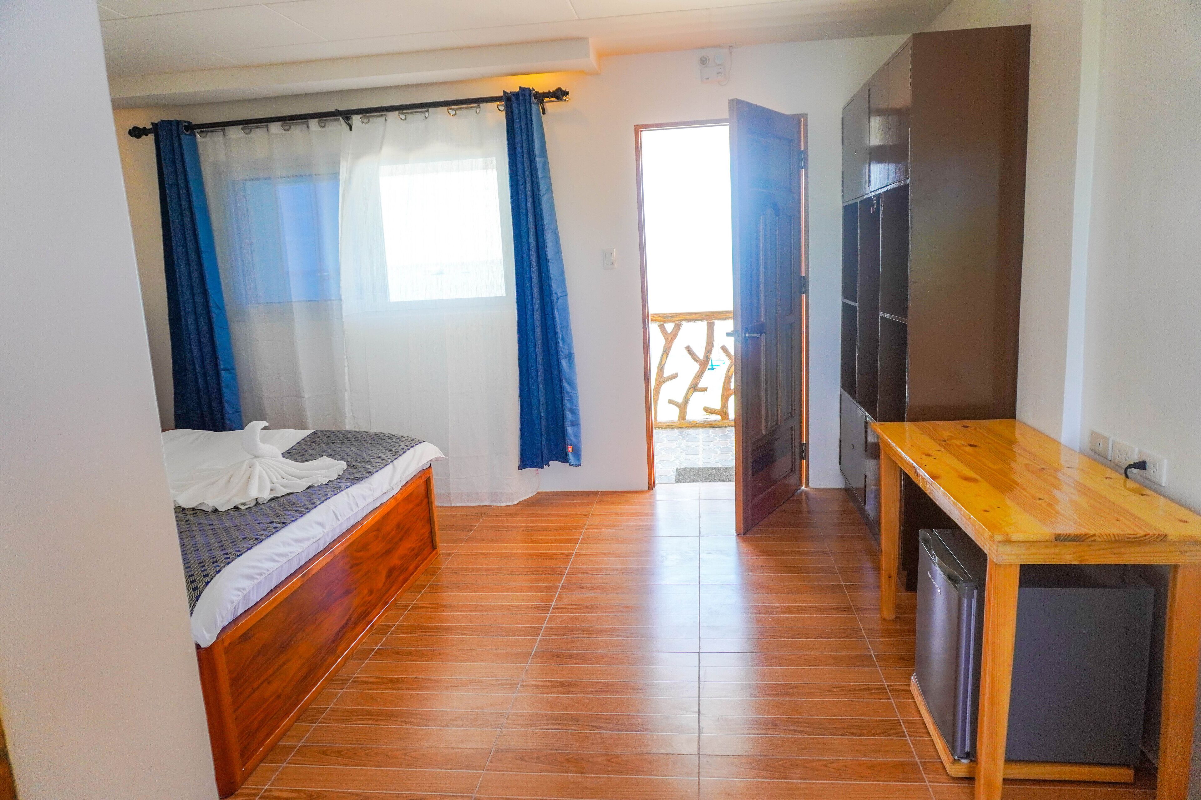 A bedroom with a wooden bed, a small wooden desk, a TV, and a balcony with a railing.