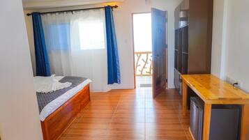 A bedroom with a wooden bed, a small wooden desk, a TV, and a balcony with a railing.