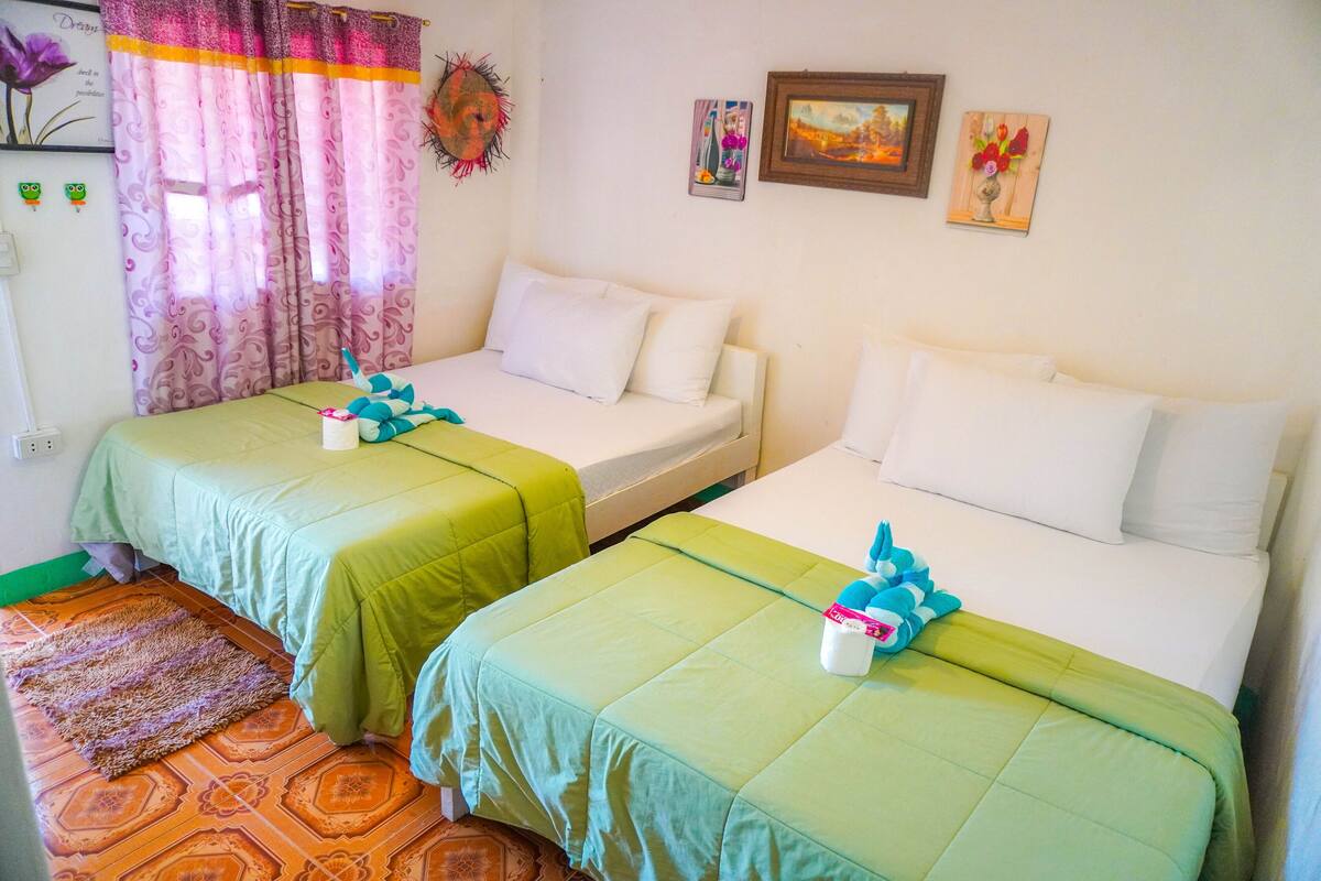 Ein Zimmer mit zwei Einzelbetten, jeweils mit einer grünen Tagesdecke und weißen Kissen. Auf dem Boden liegt ein gemustertes Teppich, und ein Vorhang mit floralem Muster ist teilweise zurückgezogen.
