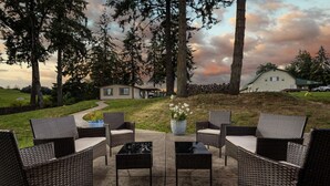 An outdoor seating area with wicker furniture and a central table, surrounded by trees and a grassy hill with a house in the background. - Single Level Ranch Layout, Huge Patio, Surrounded by Vineyards, Free Tastings, Foosball and Fire-Pit (Dayton)