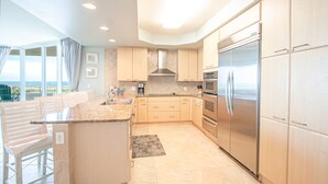 A modern kitchen with light wood cabinets, stainless steel appliances, and a large island with seating. - Beautiful Spacious Condo with Wraparound Water and Pool Views with Complimentary Boat Slip! (Orange Beach)