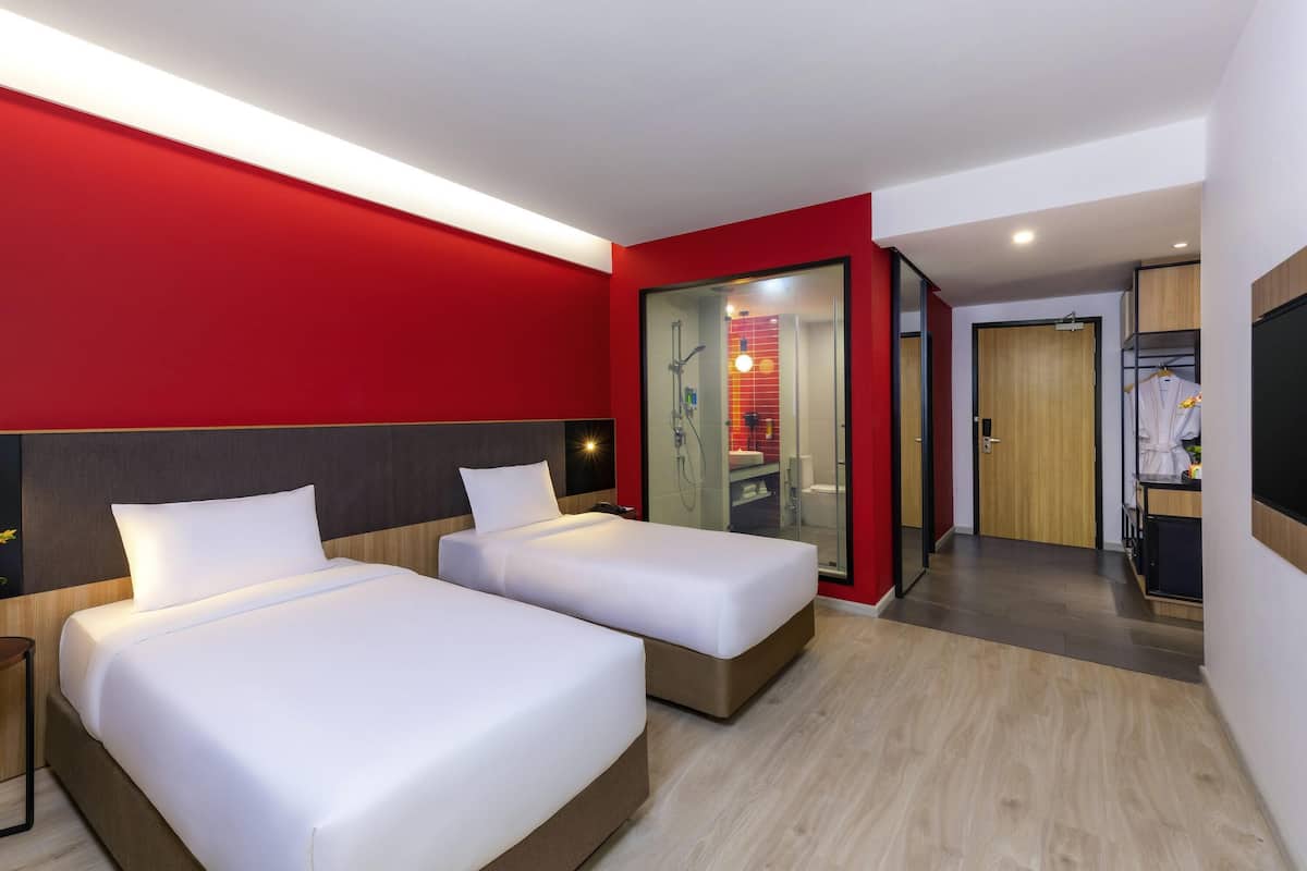 Ein Hotelzimmer mit zwei Betten, einer roten Akzentwand, einer behinderten Duschkabine aus Glas, einem Holzschrank und einem an der Wand montierten Fernseher.