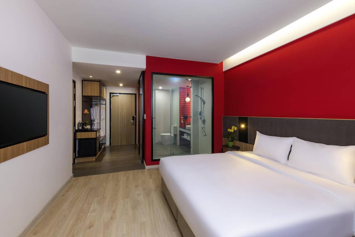 Ein Hotelzimmer mit einem großen Bett, einem an der Wand montierten Fernseher, einer roten Akzentwand und einer Duschkabine aus Glas.