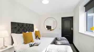 Une chambre moderne avec une tête de lit capitonnée, un miroir rond, un lit avec du linge de lit blanc et des coussins jaunes, une table de chevet avec une lampe et une fenêtre donnant sur un beau paysage.