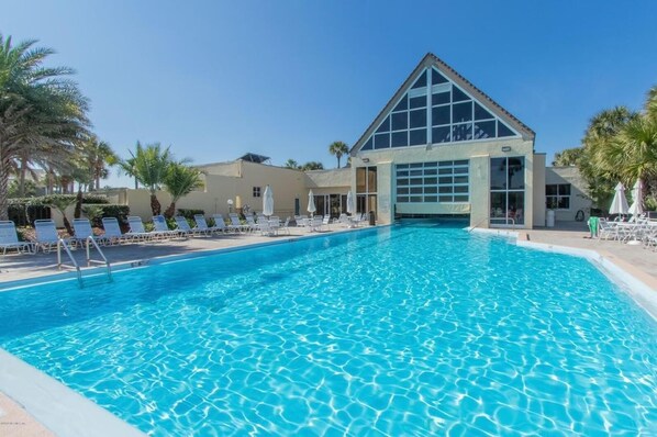 Una piscina con sillas de descanso y sombrillas, un edificio de techo triangular y palmeras.