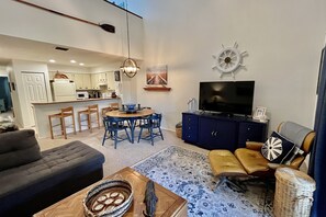 Uma sala de estar moderna com um sofá, uma mesa de jantar com cadeiras, uma televisão e uma cozinha ao fundo.