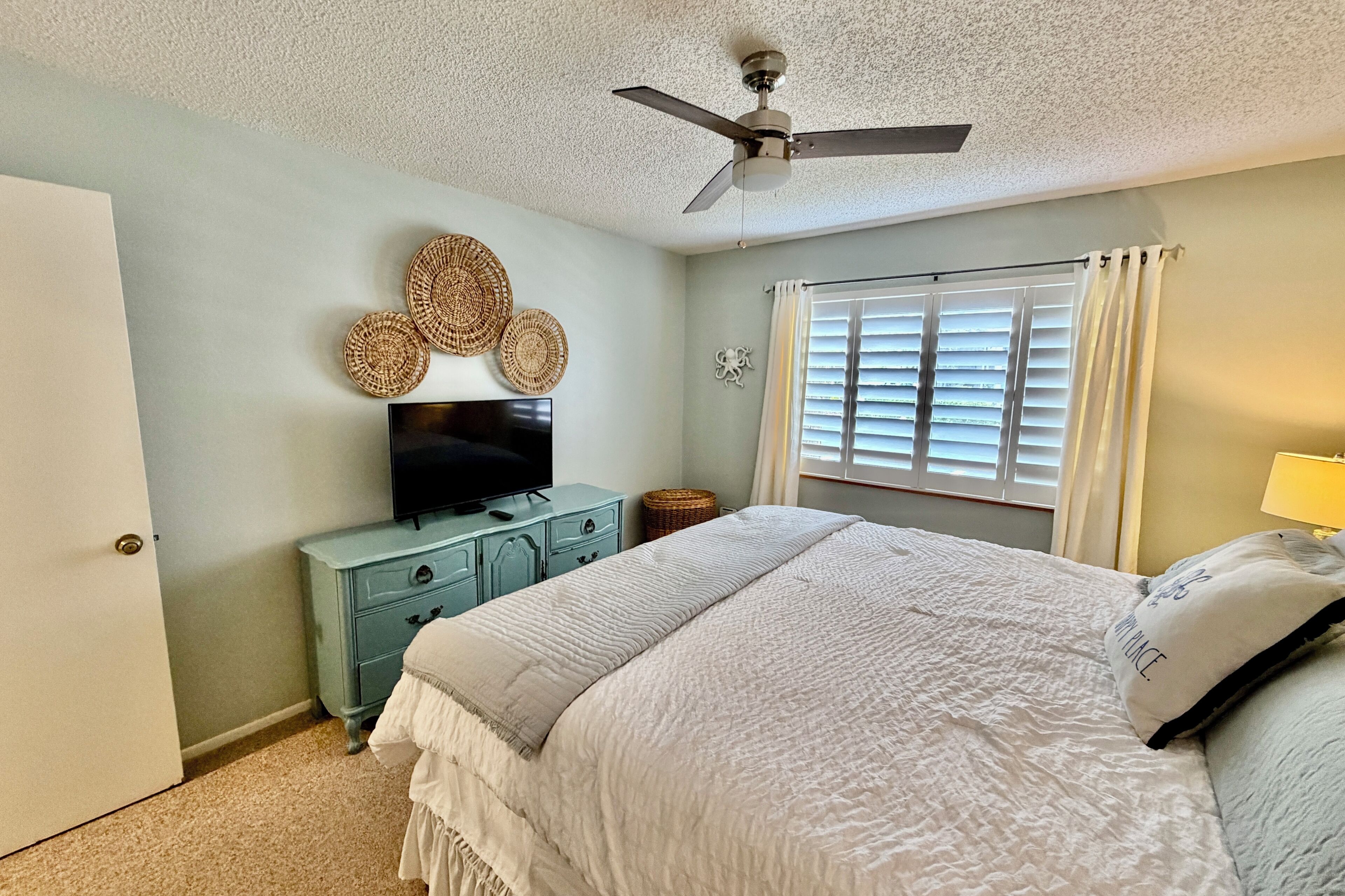A bedroom with a bed, a television on a dresser, a ceiling fan, and a window with blinds.