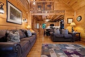 A cozy cabin interior with wooden walls, two leather sofas with patterned cushions, a dining area with a table and chairs, and a kitchen with modern appliances.