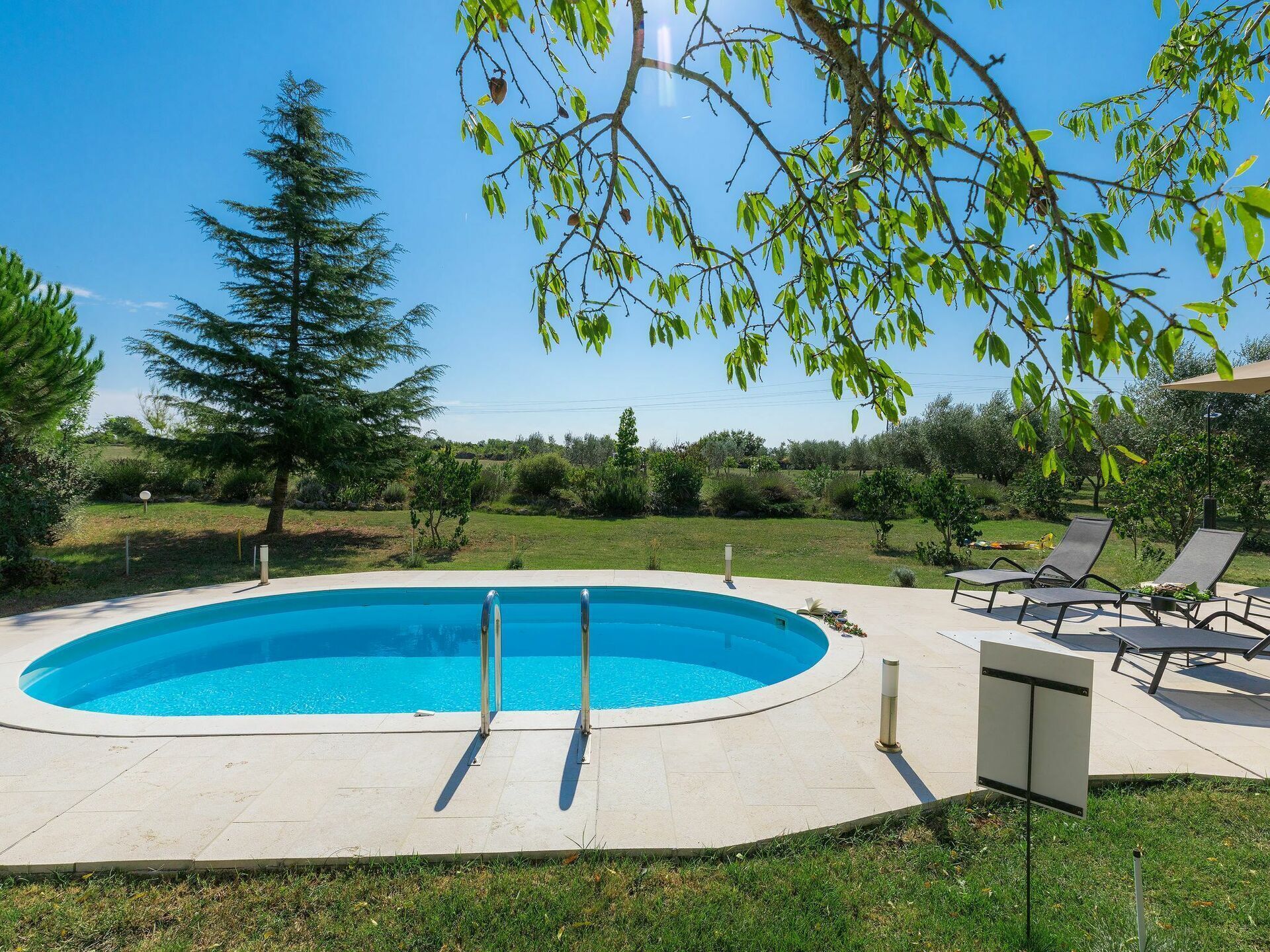 Una piscina circular con dos escaleras, rodeada por un solárium empedrado, con sillas de descanso y una zona ajardinada al fondo.