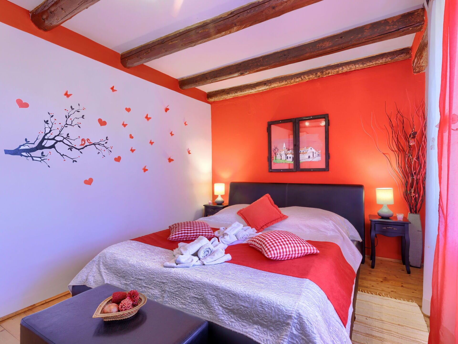 A bedroom with a bed, two bedside tables, a nightstand, a wall with a tree decal, and a window with a view of a village.