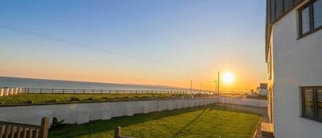 A grassy area with a wooden fence, a building with a white facade, and a view of the sea at sunset.