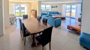 Un'ampia zona giorno con tavolo da pranzo in legno, divani blu e vista sulla spiaggia attraverso ampie finestre.