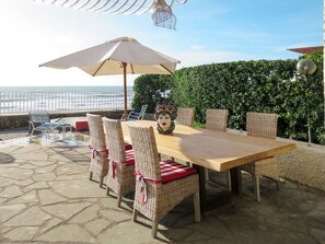 Un patio al aire libre con una mesa de madera y sillas de mimbre, un paraguas grande y vista al océano.