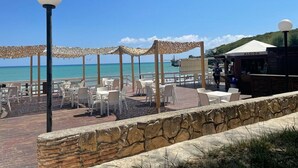Un patio frente a la playa con sillas y mesas blancas, un muro de piedra y un restaurante llamado ALGIDA al fondo.
