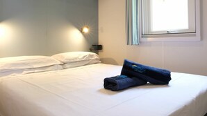 Una cama bien tendida con sábanas blancas, una toalla doblada, una lámpara de pared y una ventana con cortinas.