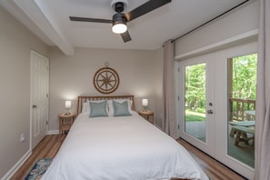 A bedroom with a large bed, a ceiling fan, a wall clock, and a sliding glass door leading to an outdoor deck.