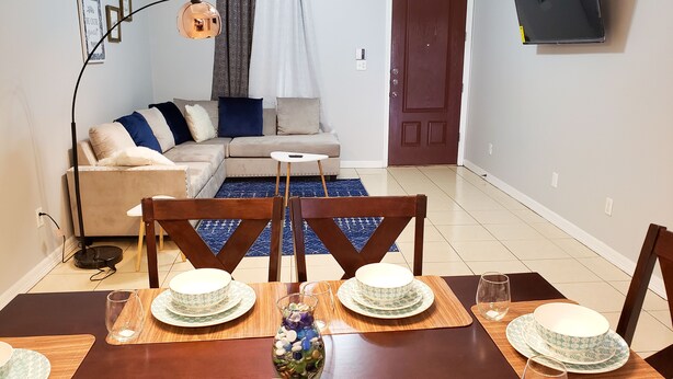 Un comedor con una mesa de madera dispuesta para cuatro personas, un sofá, un televisor montado en la pared y una puerta al fondo.