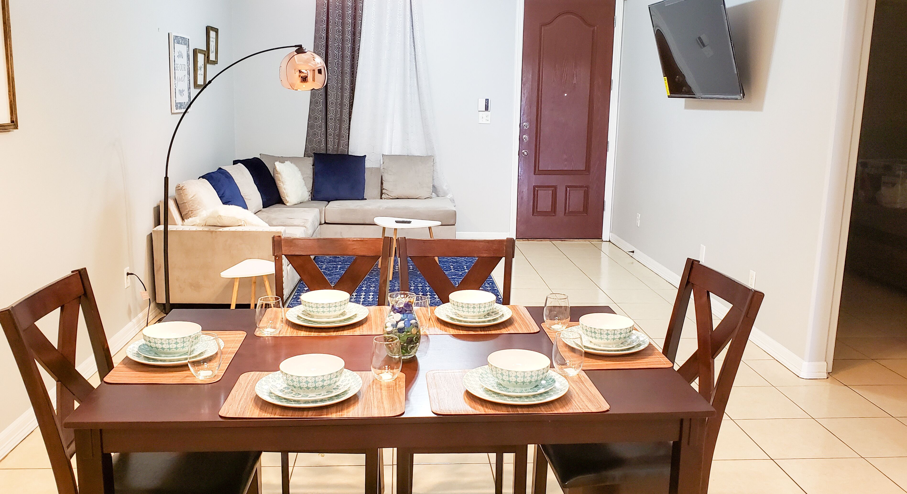 A dining area with a wooden table set for four, surrounded by wooden chairs. A sofa and a television are visible in the background.