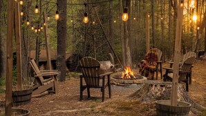 A forest campsite with a fire pit, wooden chairs, and string lights.