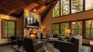 A cozy living room with a stone fireplace, a flat-screen TV, and large windows offering a forest view.