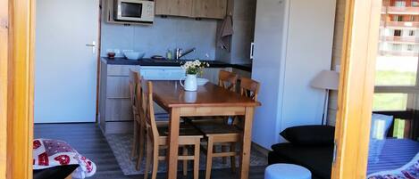 A compact kitchen with wooden cabinets, a microwave, and a refrigerator. There is a dining table with chairs and a small table with cups and saucers.