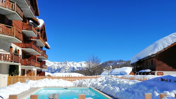 A hotel with wooden balconies, a pool surrounded by snow, and mountains in the background. - SOUTH ALPES APARTMENT PUY SAINT VINCENT 1800 M Ski resort summer / winter (Puy-Saint-Vincent)