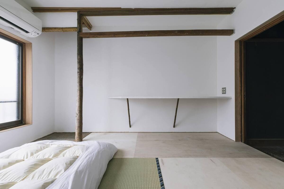 A minimalist room with a bed, a wooden beam ceiling, a white wall, and a window.